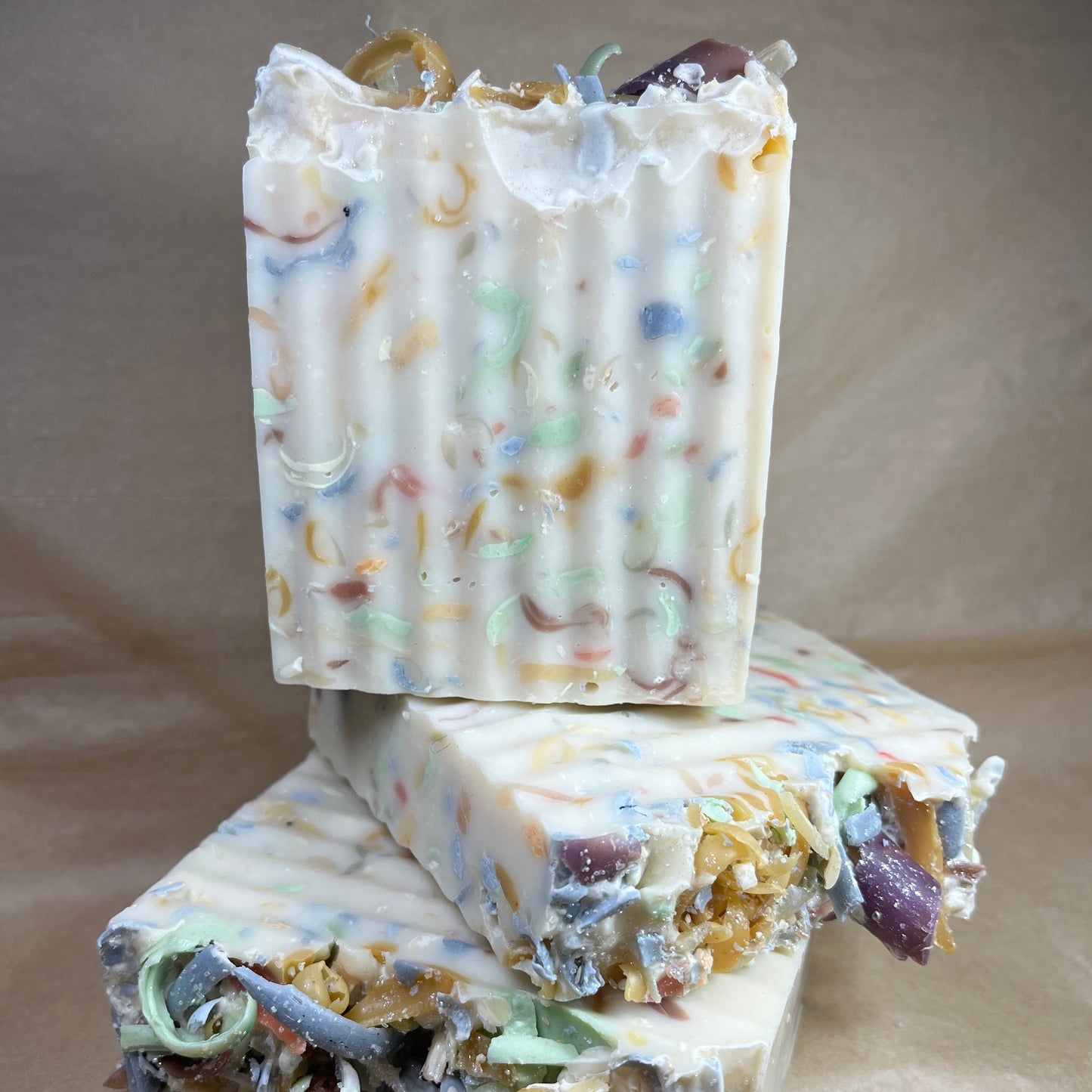 A bar of handmade soap with a white and multicolored confetti-like topping, partially melted to reveal the layers of colorful ingredients, placed on a brown surface.
