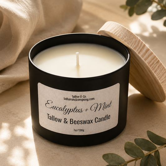 Candle in a black container with a label, surrounded by eucalyptus leaves on a beige surface.
